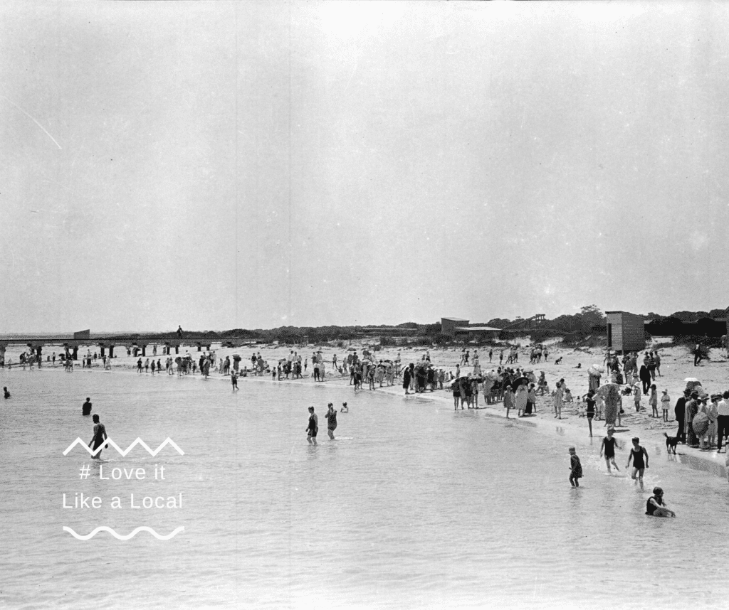 Old black-and-white photo of a crowd of people enjoying the beach by the jetty